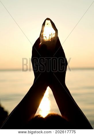 Young woman's hands holding clear quartz point in front of the lake sunrise