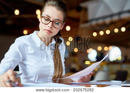 Concentrated young white collar worker wrapped up in paperwork while sitting at spacious coffeehouse, waist-up portrait shot