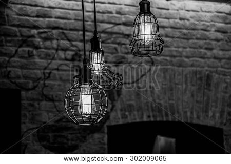 Black And White Photography Of Wire Lamps Hanged From Ceiling On Old Brick Wall Background. Glowing 