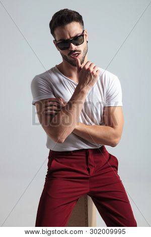Sensual young man touching his lips and looking to the camera while wearing a white t-shirt and sitting on gray studio background