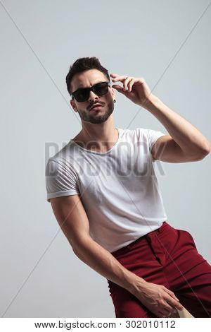 Tough guy about to take off his sunglasses while wearing a white t-shirt and red pants, sitting on white studio background