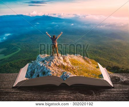 Young Hiker Man Standing On A Cliff Above The Mountain Valley On The Pages Of An Open Magical Book. 