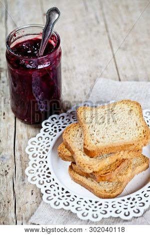 Homemade cherry jam in glassjar and fresh toasted cereal bread slices stack on white  plate closeup on linen napkin on rustic wooden table background. Sweet food for breakfast.