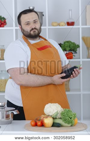 Portrait Of Guy Wearing White Skirt And Orange Apron Posing With Aubergine At Kitchen. Male Cooking 