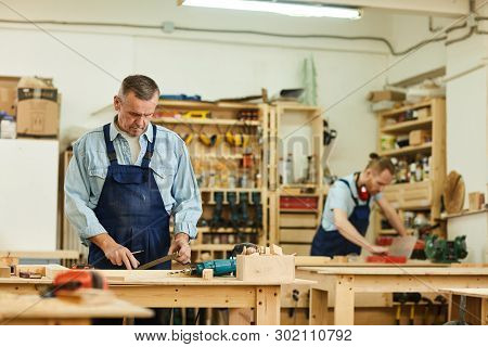 Portrait Of Two Carpenters Working With Wood Standing At Tables In Workshop, Focus On Senior Carpent