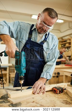 Portrait Of Mature Carpenter Drilling Wood While Working At Table In Joinery, Copy Space
