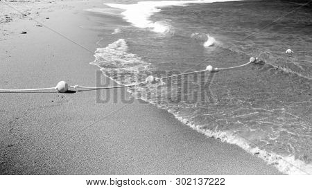 Black And White Image Of Life Saving Buoy Line On The Rope Lying On The Sandy Sea Beach And Flotaing