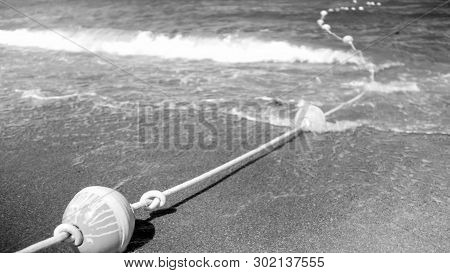 Black And White Image Of Life Saving Buoy Line On The Rope Lying On The Sandy Sea Beach And Flotaing