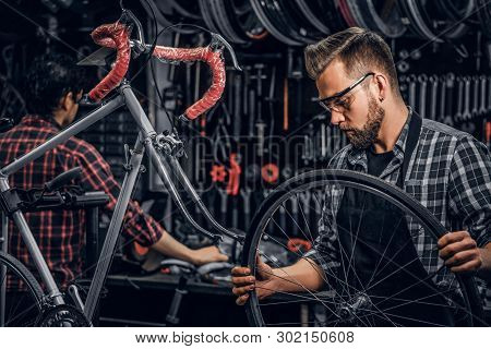 Focused Attractive Man In Glasses Is Chainging Wheel For Bicycle At Busy Workshop.