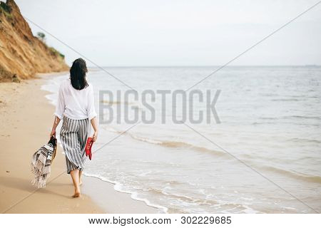 Stylish Hipster Girl Walking Barefoot On Beach, Holding Bag And Shoes In Hand. Summer Vacation. Spac