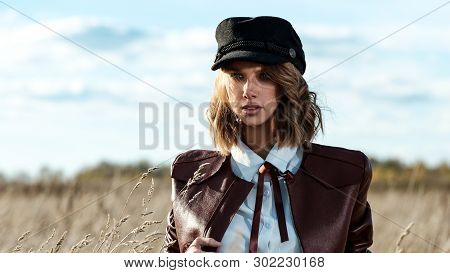 Portrait Of Attractive Female Model In Leather Jacket And Fashionable Cap Looking At Camera Against 