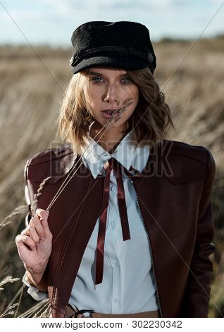 Portrait Of Attractive Female Model In Leather Jacket And Fashionable Cap Looking At Camera Against 
