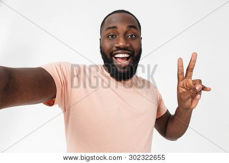 Portrait of a happy african man wearing t-shirt standing isolated over white background, taking selfie, peace gesture