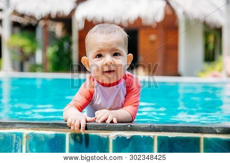 A Cute And Happy Little Baby Holds To The Side Of The Tropical Pool And Looks To The Camera And Laug