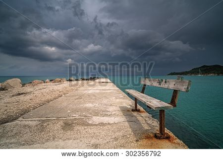 Empty Wooden Bench On A Concrete Pier With Stormy Dramatic Clouds Accumulating Over Alykes Bay In Za
