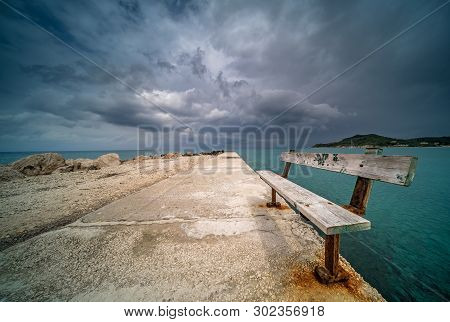 Empty Wooden Bench On A Concrete Pier With Stormy Dramatic Clouds Accumulating Over Alykes Bay In Za