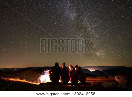 Night Summer Camping In Mountains. Back View Group Of Four Hikers Sitting On A Bench Made Of Logs To