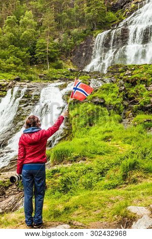 Tourist Woman With Norwegian Flag At Svandalsfossen In Norway, Powerful Waterfall In Norwegian Mount