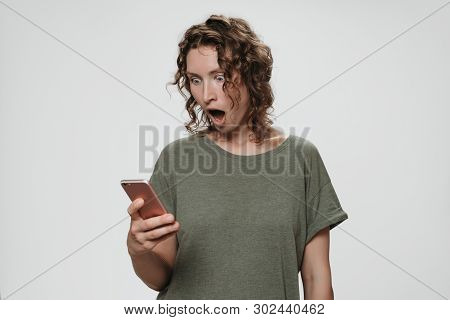 Puzzled Stupefied Curly Hair Woman Opens Eyes And Mouth Widely Holding Smartphone Looks With Terrifi