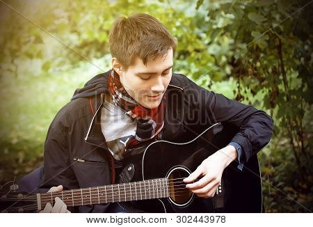 A Young Guy In A Black Windbreaker And A Plaid Scarf Plays A Black Acoustic Guitar, Sitting In The P
