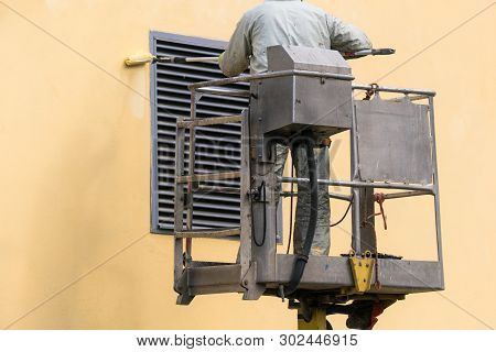 Man On A Lifting Platform Painting The Building Wall With A Roller Exterior Outdoors. Worker On A La