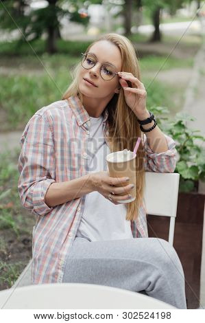 Blonde In A Coffee Shop At A Table On The Street. Paper Cup Of Coffee In Hand. Coffe To Go