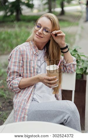 Blonde In A Coffee Shop At A Table On The Street. Paper Cup Of Coffee In Hand. Coffe To Go