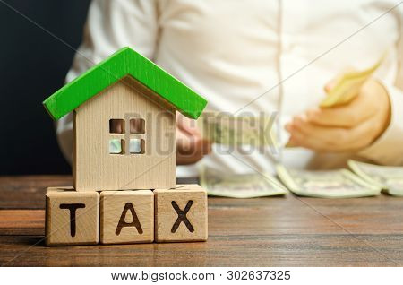 Wooden Blocks With The Word Tax, House With Money In The Hands Of A Businessman. The Concept Of Payi