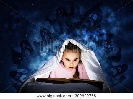 Engrossed Little Girl Reading Book In Bed After Bedtime. Kid In Pajamas With Flashlight Hiding Under