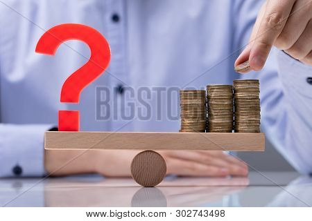 Hand Placing Coin On Top Of The Stack Coins And Question Mark Sign Over Seesaw