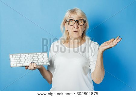 Old Lady With Glasses Holds A Keyboard On A Blue Background. Internet Concept For Old People, New Te