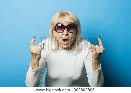 Old Woman Wearing Glasses With A Surprised Face Makes A Gesture With Her Hands Against A Blue Backgr