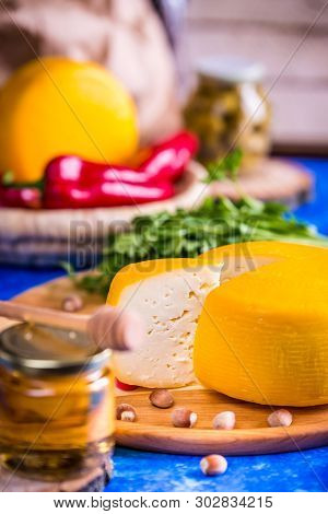 Cheese Wheel On A Wooden Board Lies On The Table With Food And Fresh Herbs. Blurred Background.