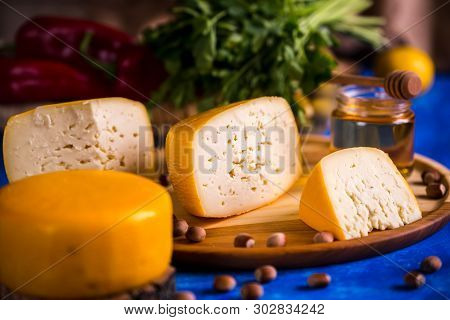 Cheese Wheel On A Wooden Board Lies On The Table With Food And Fresh Herbs. Blurred Background.