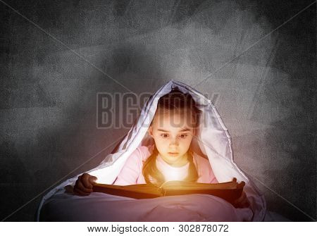 Engrossed Little Girl Reading Book In Bed Before Going To Sleep. Pretty Kid With Flashlight Hiding U