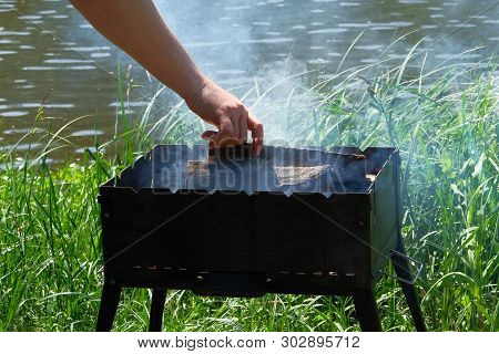 Grill With Fuel And Charcoal. Hand Is Adding Wood Into The Fire. Barbecue Near River. Sun.
