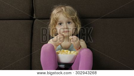 Beautiful Little Blonde Girl Sitting On A Sofa At Home And Eating Corn Puffs. Cute Child Taste Puffc