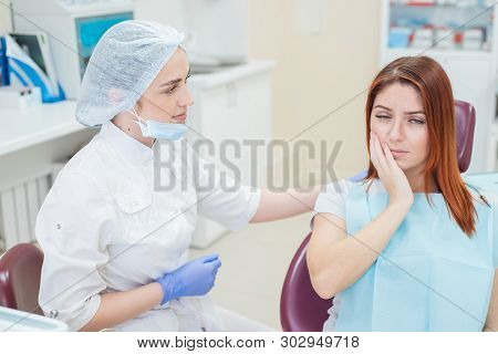 Young Beautiful Female Dentist Helps Patient With Toothache. A Red-haired Woman Sits In A Chair At T