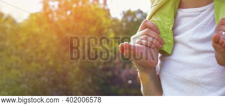 A Man With A Child On His Shoulders. Close-up Of Barefoot Little Leg And Hand Of Father On The Backg