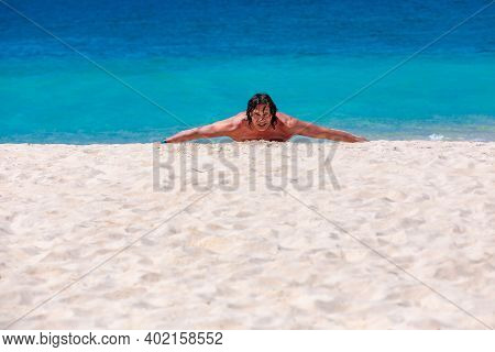 A Young Man With Long Black Hair Is Lying On The Sand By The Sea, His Arms Spread Wide. Blue Waves, 