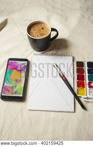 Set Of Watercolor Paints, Scetchbook, Paintbrush And A Cup Of Coffee For Breakfast.