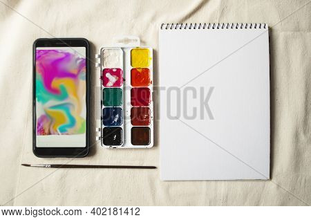 Set Of Watercolor Paints, Scetchbook, Paintbrush And A Smartphone With Picture For Reference.
