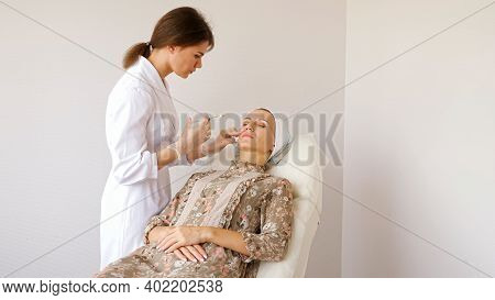 Professional Beauty Salon Worker In White Coat And Sterile Gloves Injects Young Woman Cheek On Large