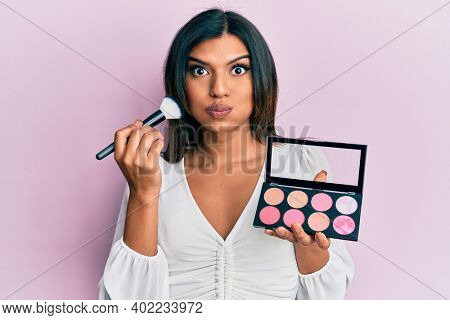 Young latin transsexual transgender woman holding makeup brush and blush palette puffing cheeks with funny face. mouth inflated with air, catching air. 