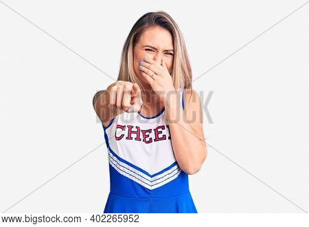 Young beautiful blonde woman wearing cheerleader uniform laughing at you, pointing finger to the camera with hand over mouth, shame expression 
