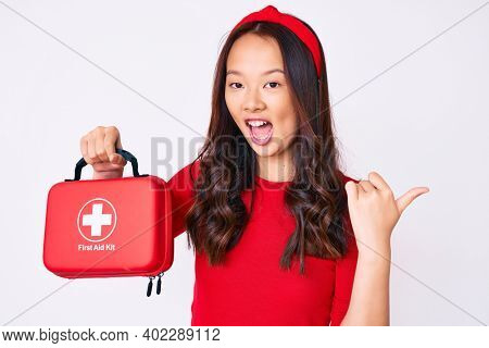 Young beautiful chinese girl holding first aid kit pointing thumb up to the side smiling happy with open mouth 