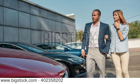 Happy Young Caucasian Pair Choosing New Car For Themselves While Walking Along Row Of Electric Autos