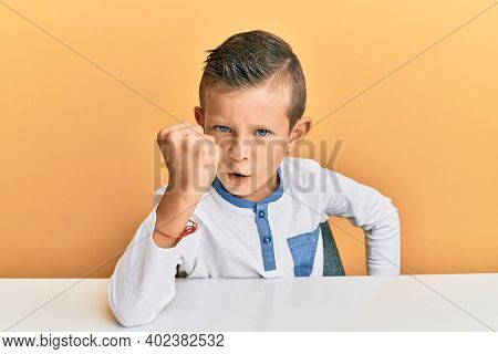 Adorable caucasian kid wearing casual clothes sitting on the table angry and mad raising fist frustrated and furious while shouting with anger. rage and aggressive concept. 