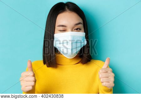 Covid-19, Social Distancing And Pandemic Concept. Cute Asian Girl In Medical Mask Winking At Camera,