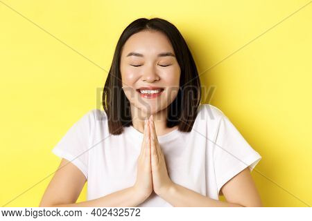 Close Up Of Hopeful Asian Girl In White T-shirt, Holding Hands In Pray, Namste Gesture And Smiling, 